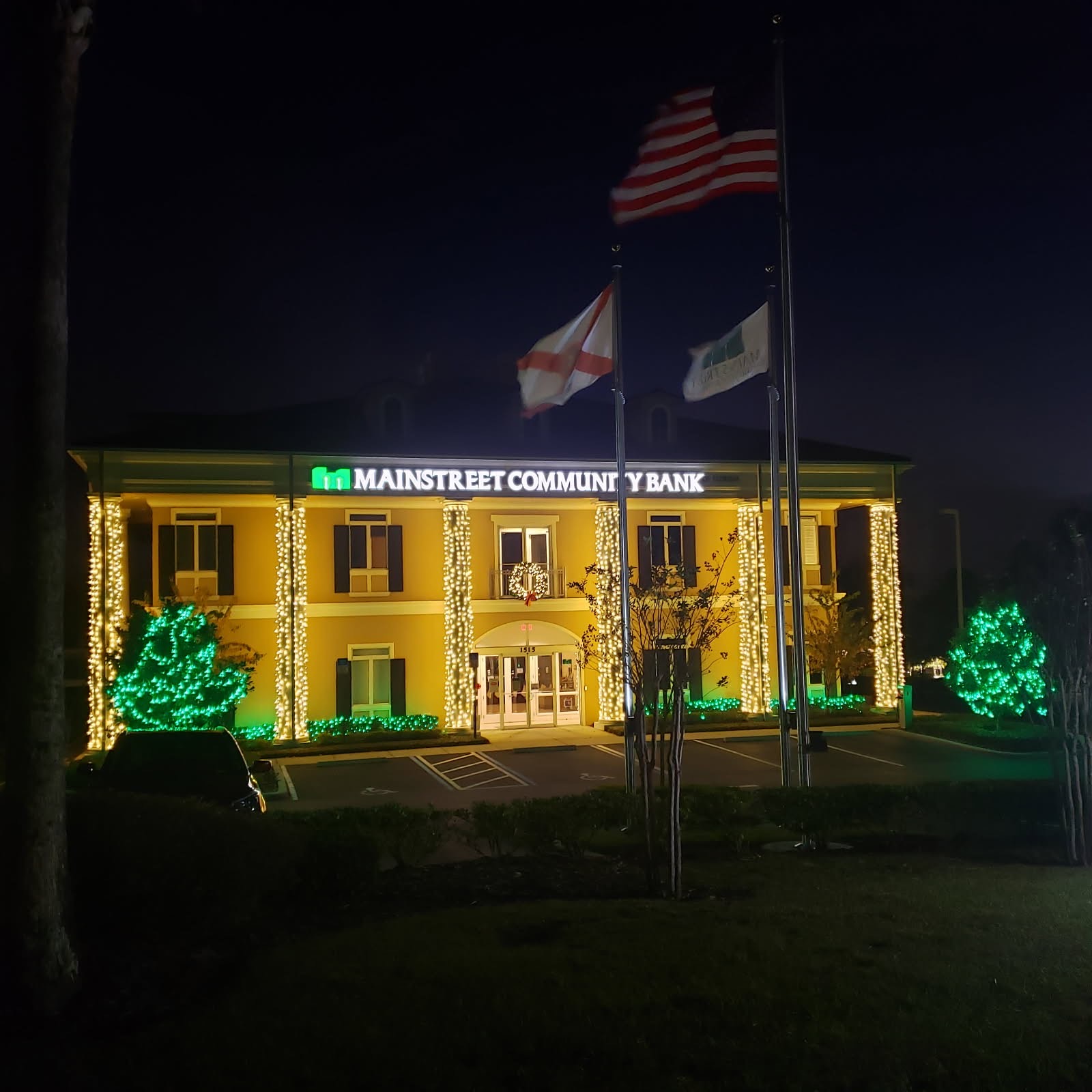 Mainstreet Community Bank at night with warm white lights wrapping the columns and green-lit trees at the entrance