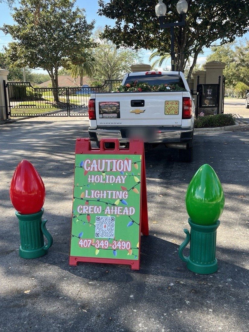 Lipham Lighting work truck behind a Caution Holiday Lighting Crew Ahead sign flanked by giant ornament props
