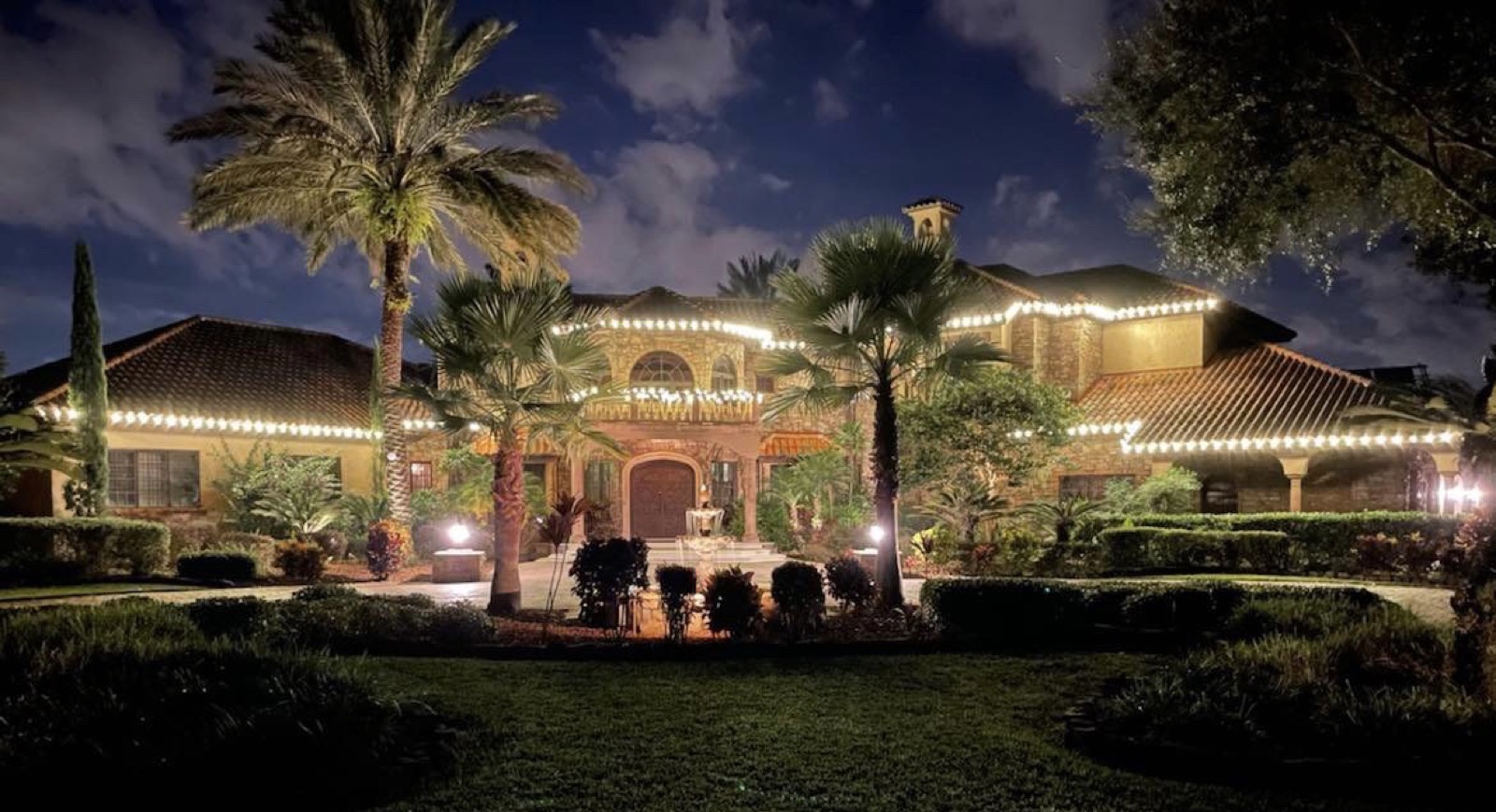 Mediterranean estate with warm white roofline lighting and uplit palms at night