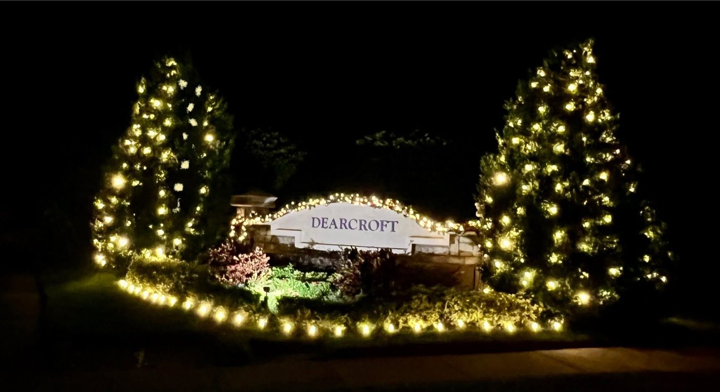 Dearcroft community monument sign with lit holiday garland and two fully lit trees flanking the entrance at night