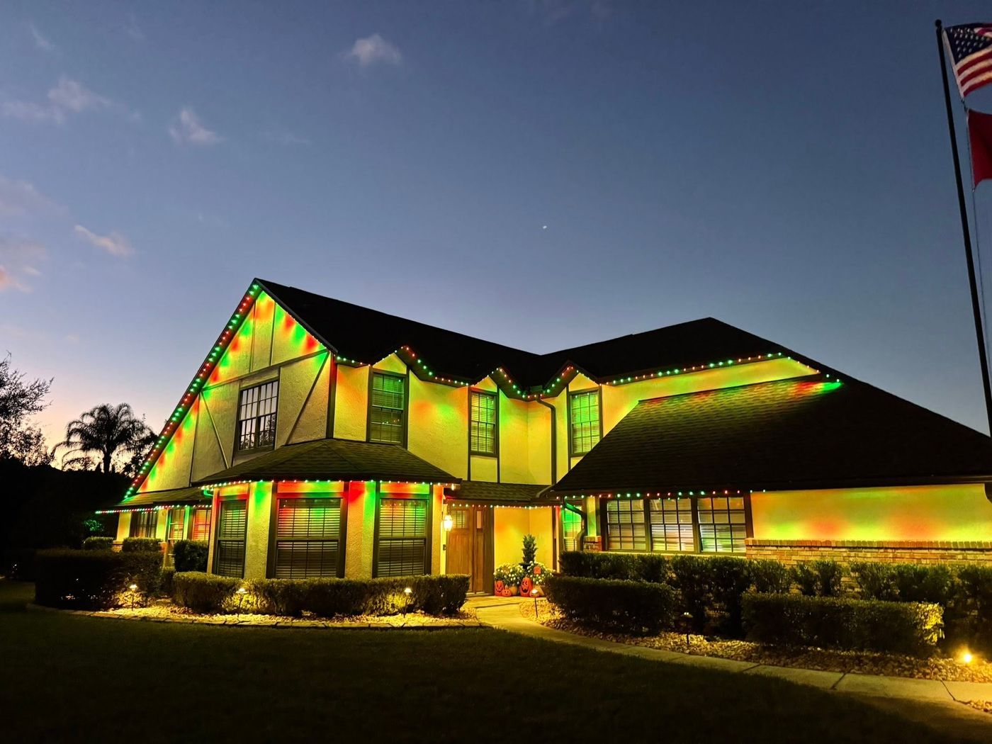 Large two-story estate with red and green holiday roofline lighting and flags at twilight