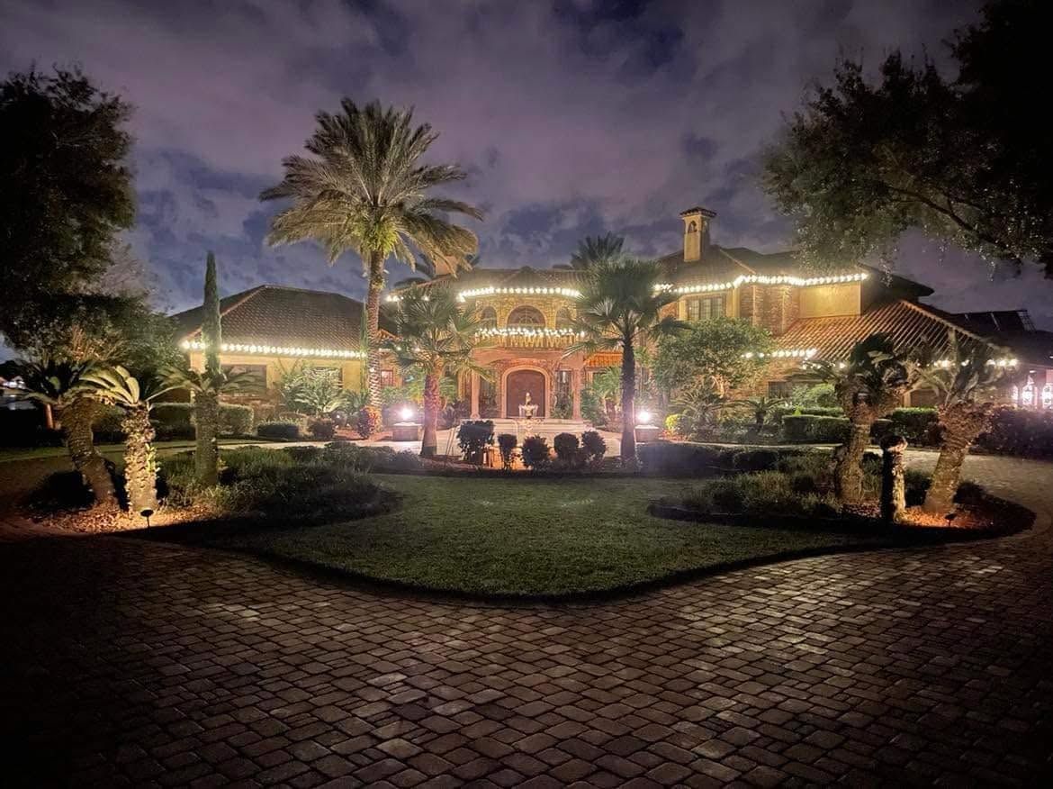 Mediterranean-style estate with warm white roofline lighting and palm tree uplighting at night