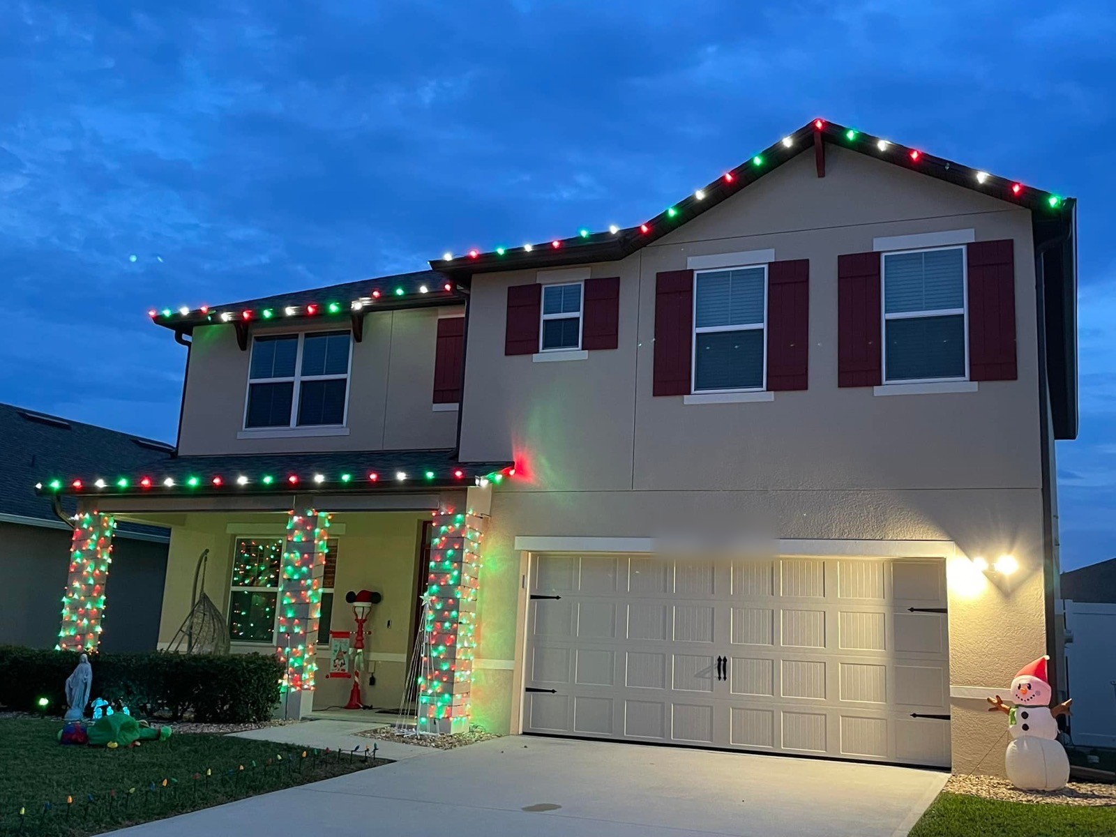Two-story home with red and green holiday roofline lighting and light-wrapped porch columns at dusk