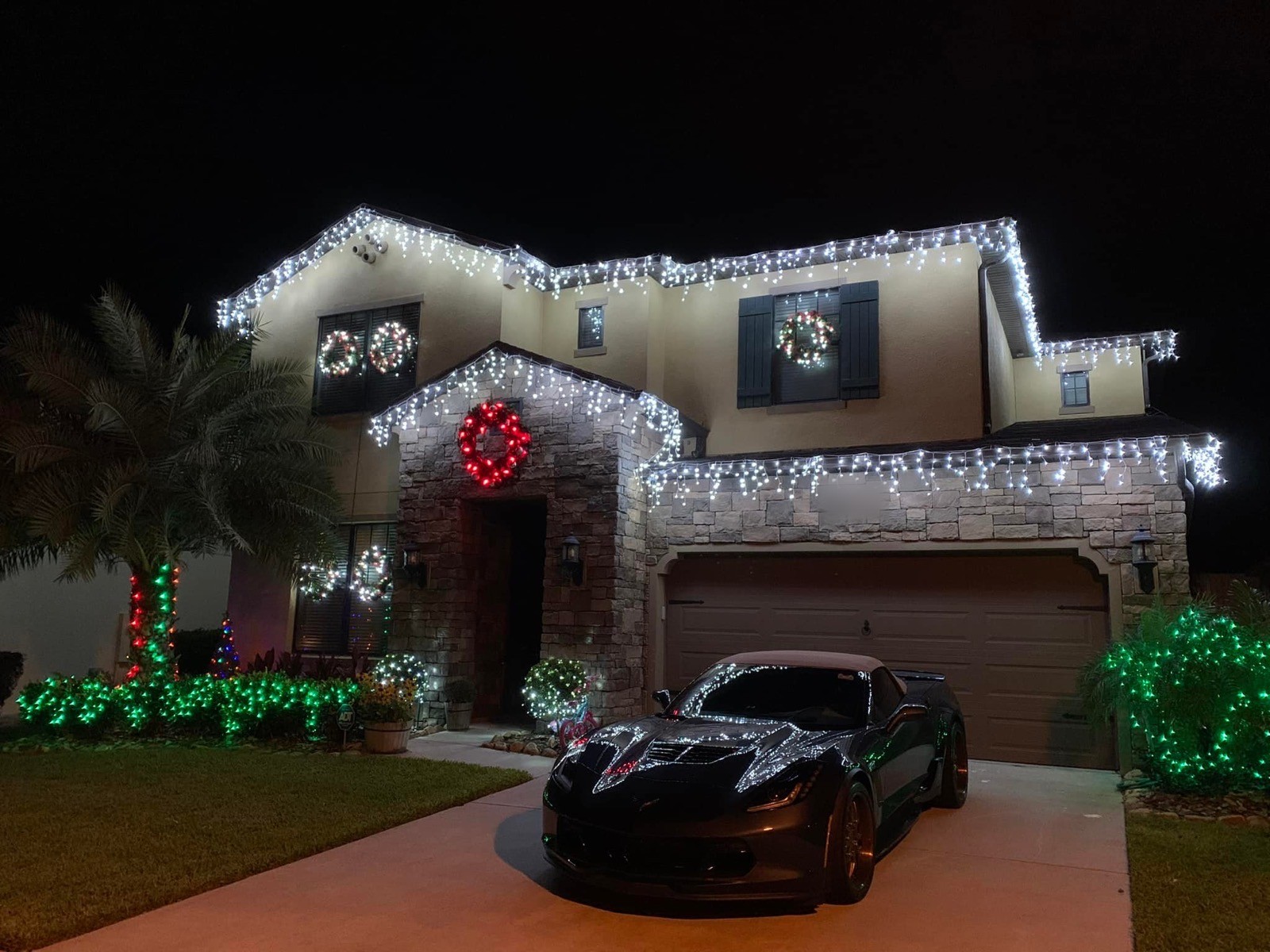 Two-story stone home draped in white icicle lights with lit wreaths and glowing green shrubs at night