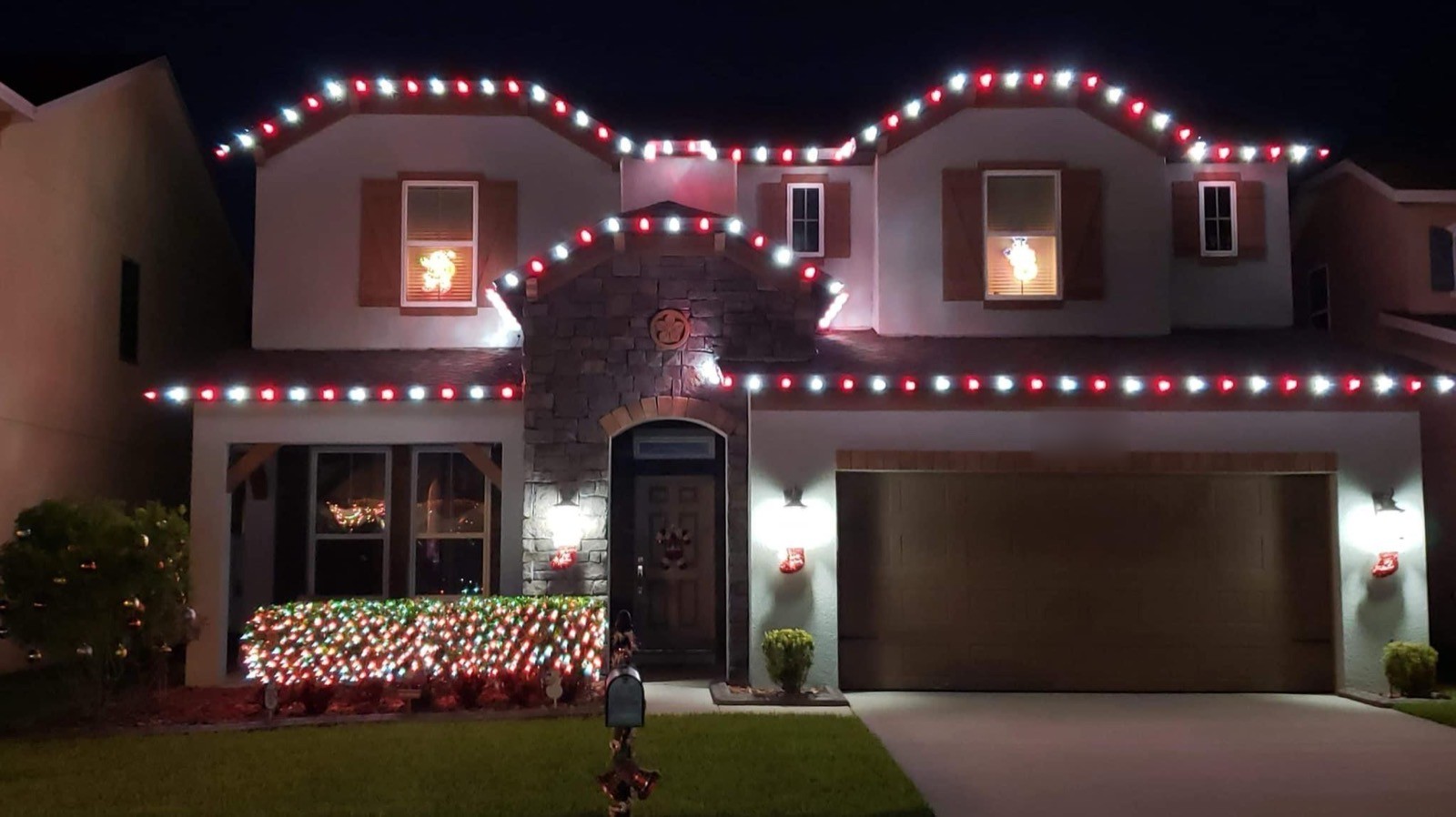 Two-story home with red and white holiday roofline lighting and a candy-striped lit hedge at night